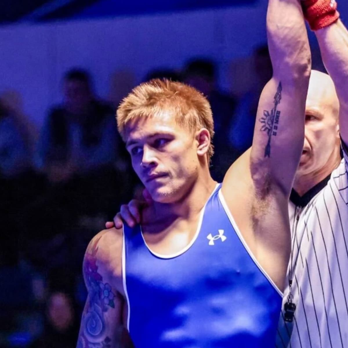 A wrestler in a blue singlet, arm raised by the referee.
